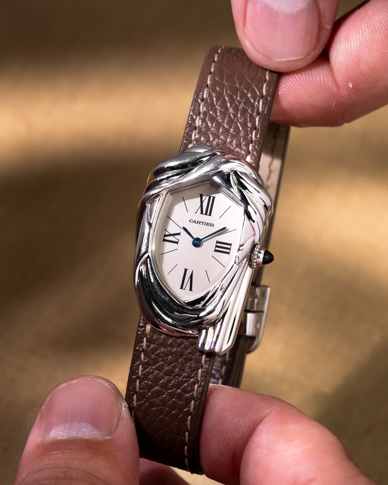 The white-gold Cheich held between two fingers by its brown strap, the woven case catching the light