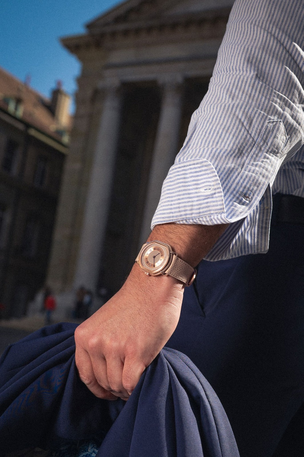 The Extra Plat Rose Gold worn on a wrist in bright sunlight, striped shirt cuff turned back, navy fabric in hand and a columned stone facade behind
