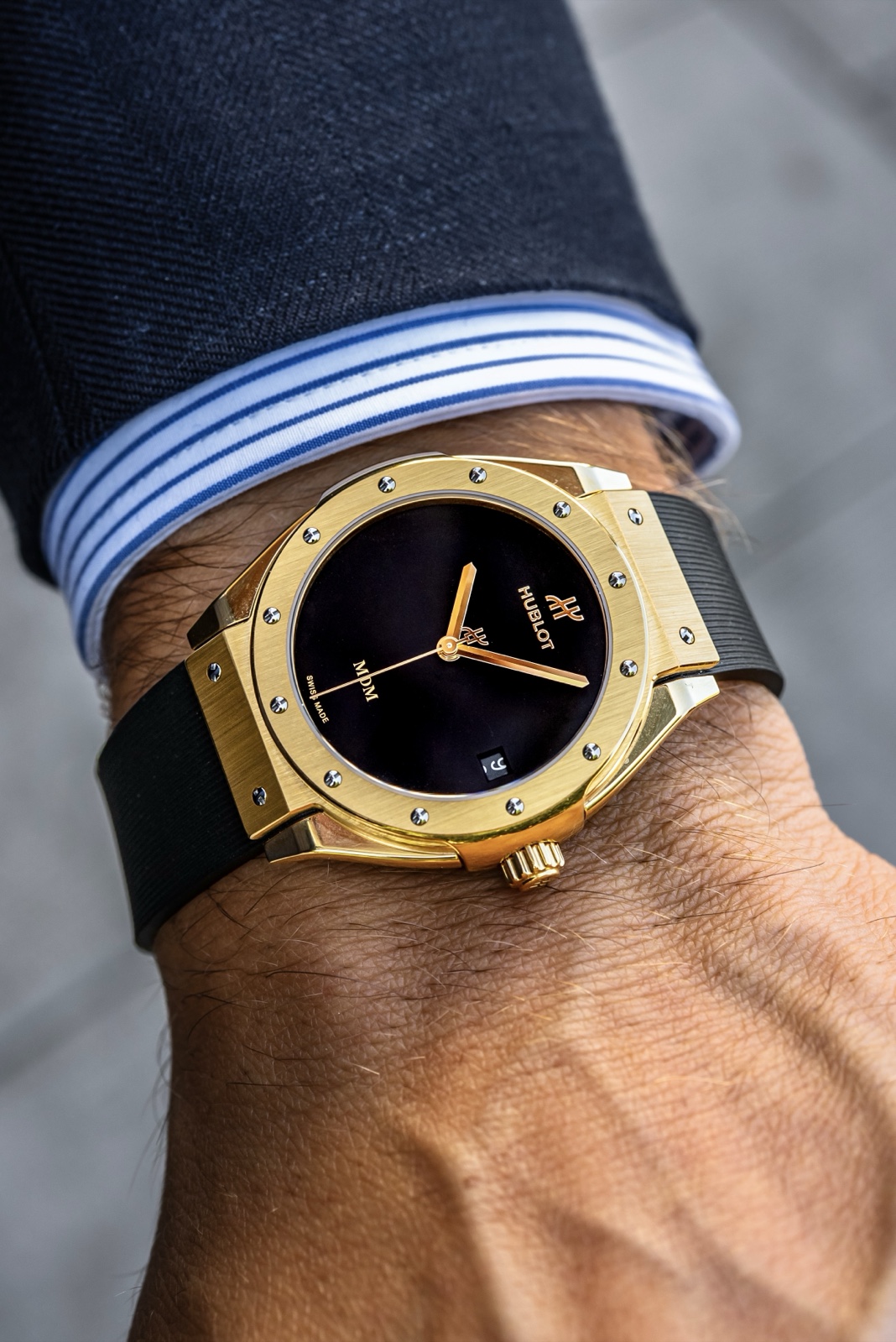 Yellow gold Hublot Classic Fusion Tributo with a black dial and ribbed rubber strap on a wrist beside a striped shirt cuff