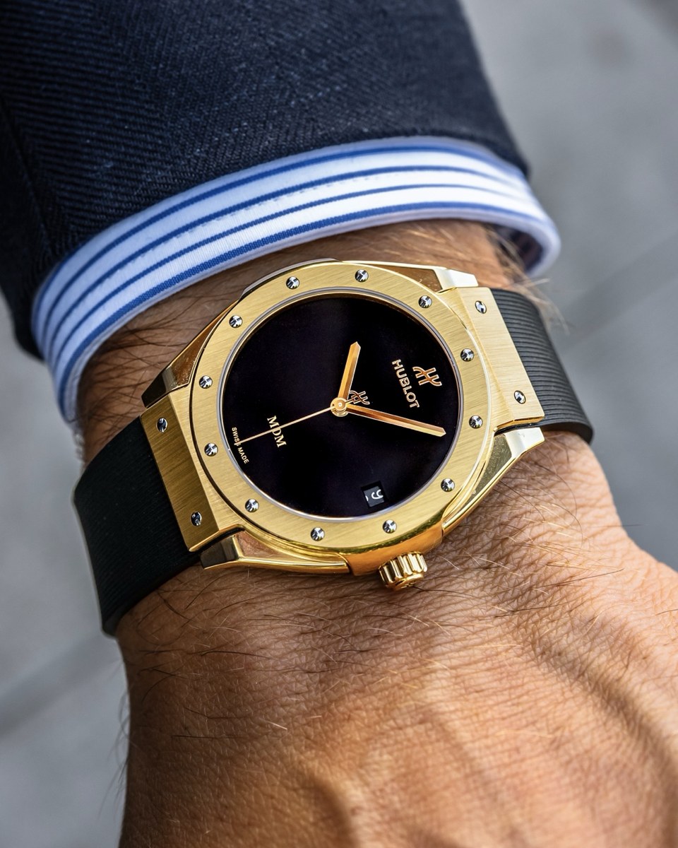 Hublot Classic Fusion Tributo in yellow gold with a black dial and black rubber strap, worn under a striped shirt cuff