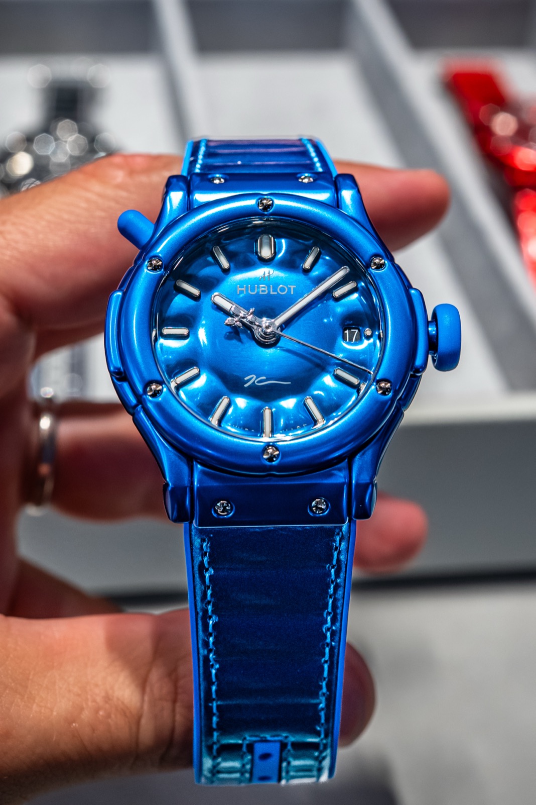 Front view of the blue Hublot watch held in one hand, with red and silver watches blurred in the tray behind it