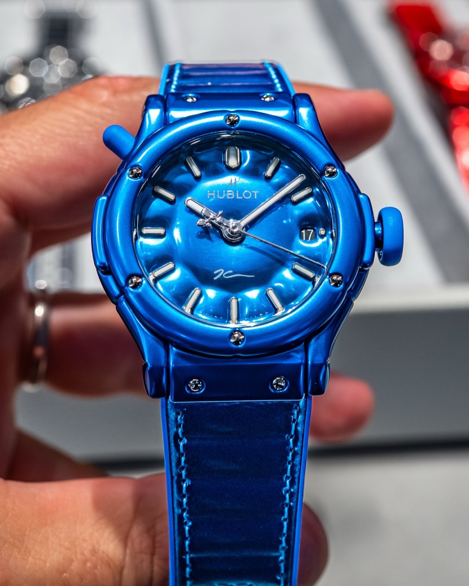 Front view of the blue Hublot watch held in one hand, with red and silver watches blurred in the tray behind it