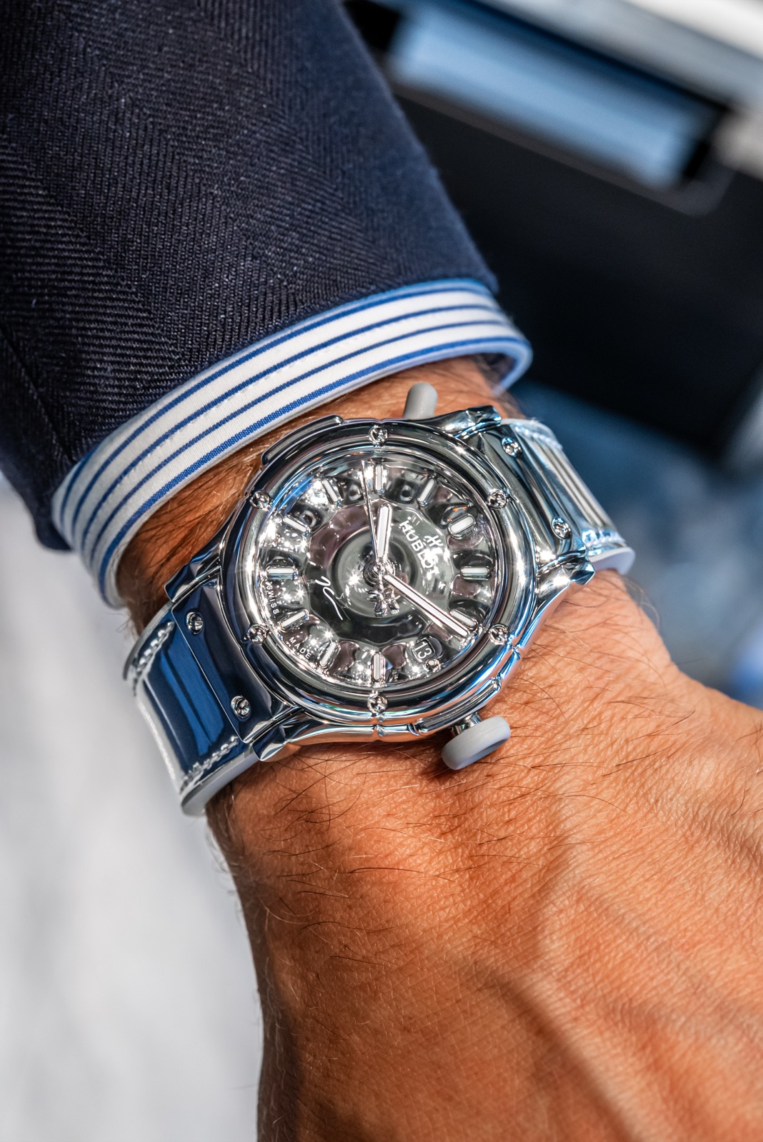 Reflective silver Hublot watch on a wrist with a dark jacket and striped cuff, photographed from above