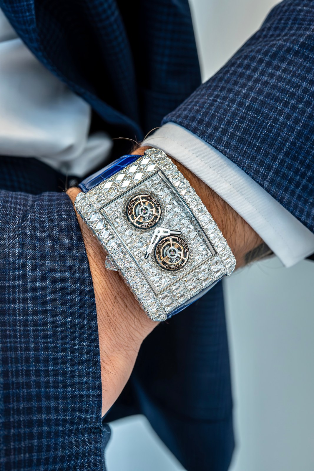 The white-diamond Billionaire Double Tourbillon Angel-Cut on a wrist under a blue checked jacket cuff, with two tourbillons visible through the set dial