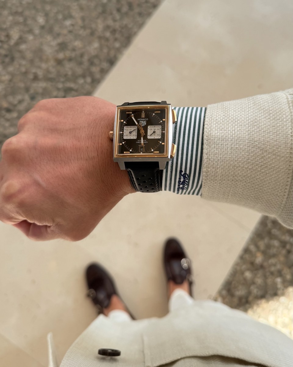 A square TAG Heuer Monaco chronograph with a dark dial and a black perforated strap on a wrist, beside a striped shirt cuff