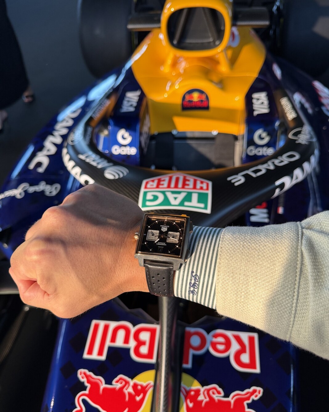 A TAG Heuer Monaco Chronograph with a black dial worn in front of an Oracle Red Bull Racing car carrying the TAG Heuer logo