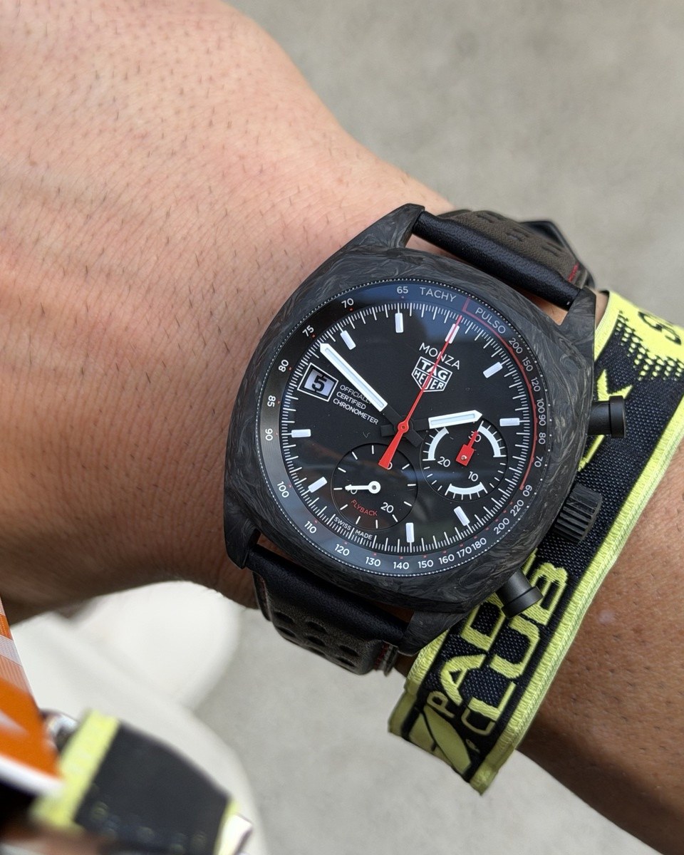 A TAG Heuer Monza chronograph with a dark, mottled case and a black leather strap on a wrist, beside a yellow Formula 1 Paddock Club wristband