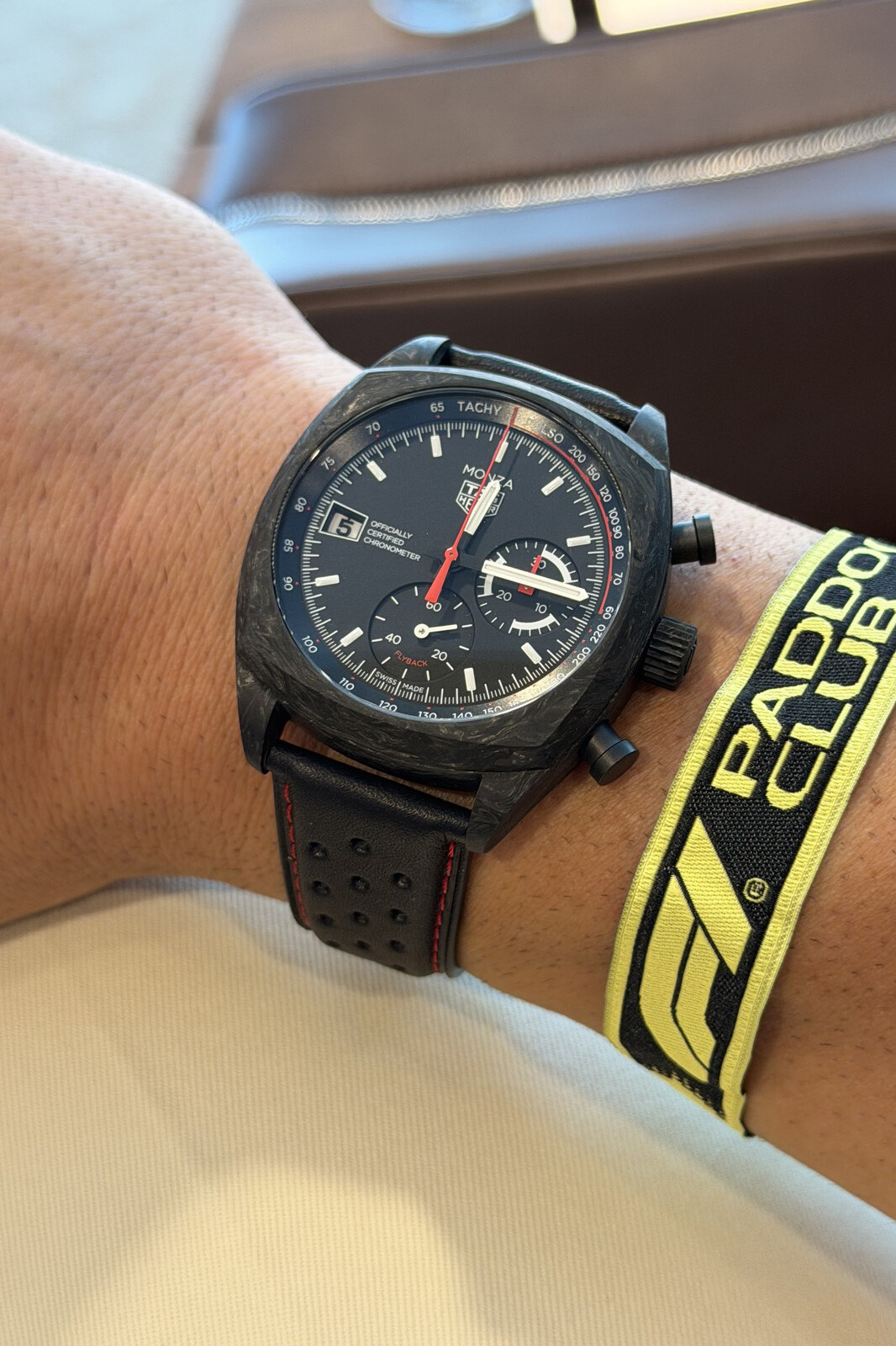 A TAG Heuer Monza chronograph with a dark, mottled case on a wrist beside a yellow Formula 1 Paddock Club wristband