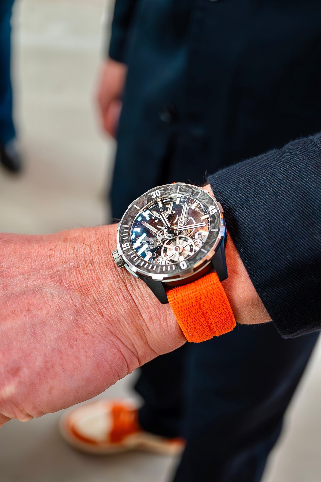 The Diver [AIR] on the orange strap on a wrist, the wearer in a dark jacket, the skeleton dial catching light